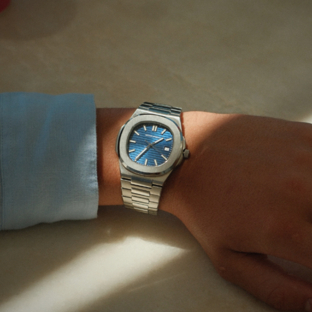 Hand wearing a silver watch with a blue face on a light surface.